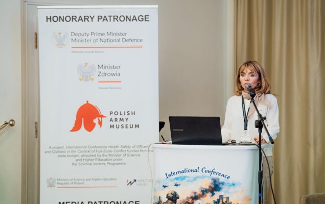 Katarzyna Gniadek przy mównicy, obok niej stoi plakat konferencyjny.