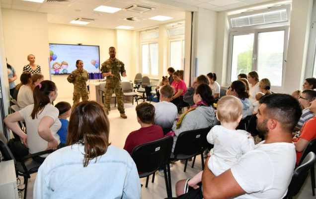 Siedzące na sali dzieci i ich opiekunowie podczas prezentacji medycznej. Przed nimi żołnierze podczas prezentacji.