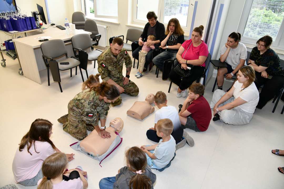 Dzieci i żołnierze siedzący na podłodze podczas pokazu pierwszej pomocy.