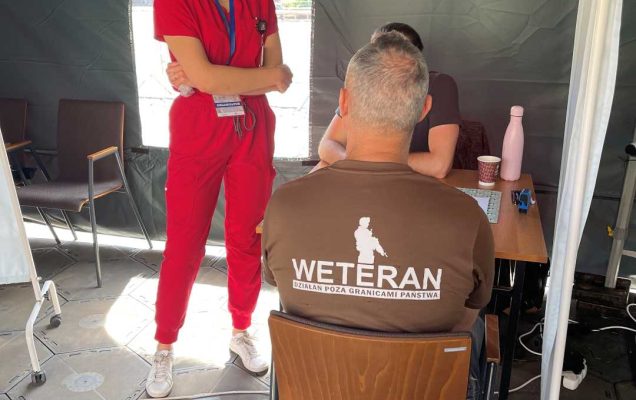 Namiot wystawowy Centrum leczenia Weterana. W środku na krzesełku siedzi mężczyzna w koszulce z napisem na plecach Weteran. Przed nim stoi pracownik Centrum leczenia Weterana.