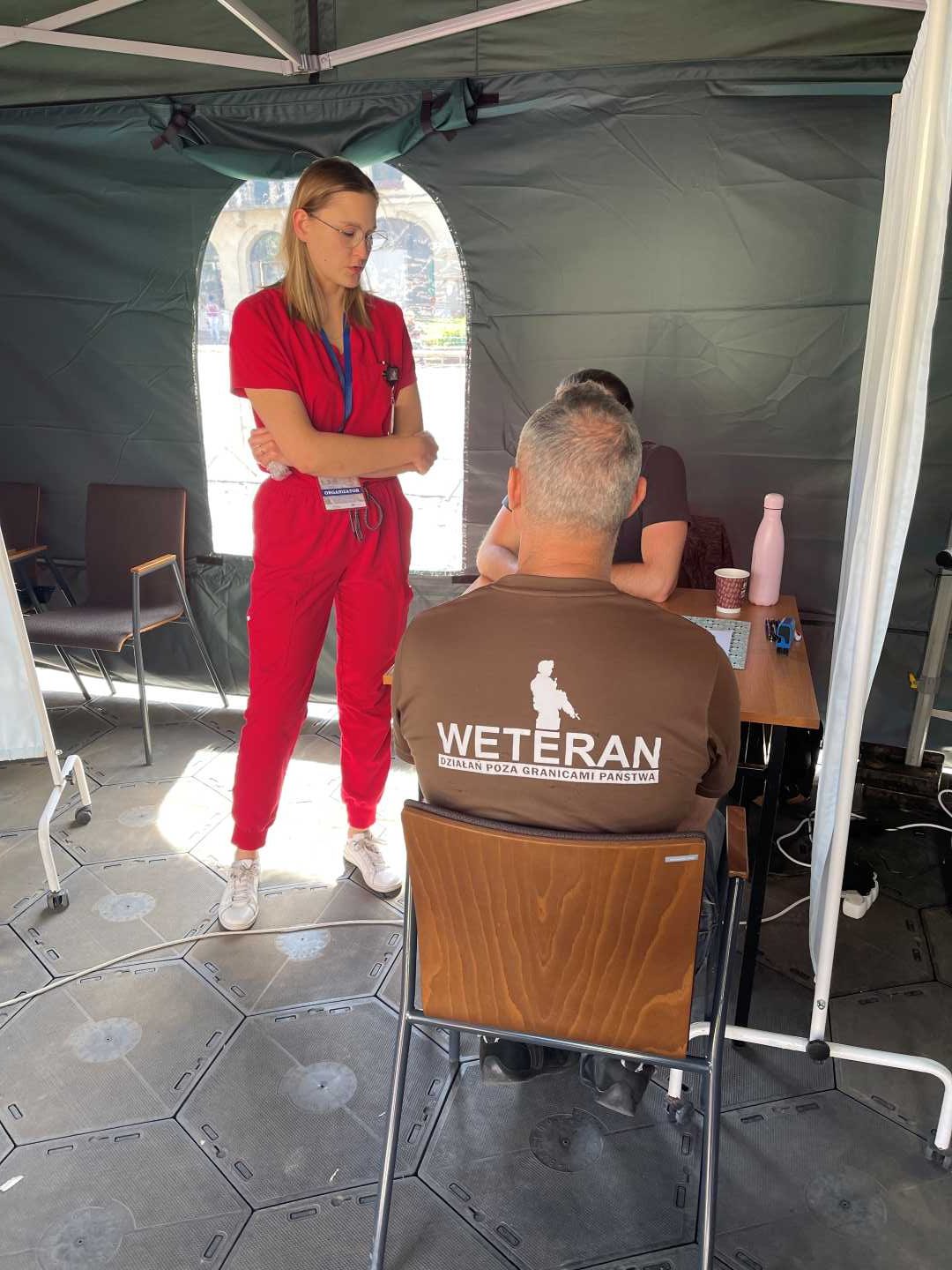 Namiot wystawowy Centrum leczenia Weterana. W środku na krzesełku siedzi mężczyzna w koszulce z napisem na plecach Weteran. Przed nim stoi pracownik Centrum leczenia Weterana.