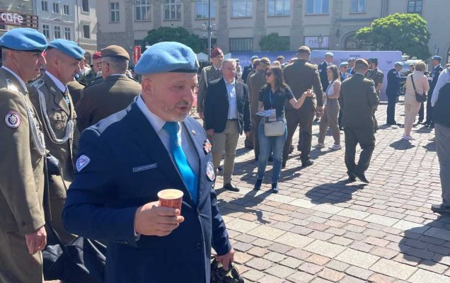 Weteran na rynku krakowskim w niebieskim berecie idzie z papierowym kubkiem z napojem w dłoni.