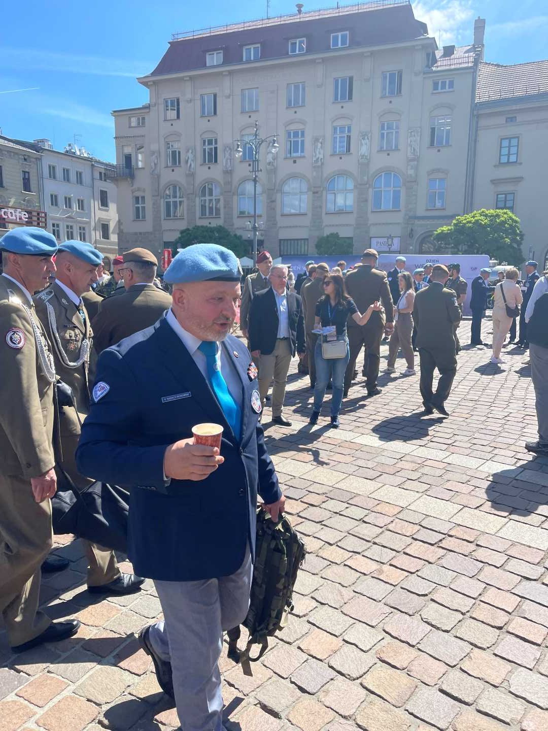 Weteran na rynku krakowskim w niebieskim berecie idzie z papierowym kubkiem z napojem w dłoni.