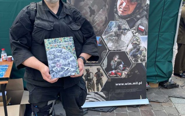 Żołnierz przy punkcie namiocie wystawienniczym Centrum Leczenia Weterana prezentuje okładkę okolicznościowej książki.