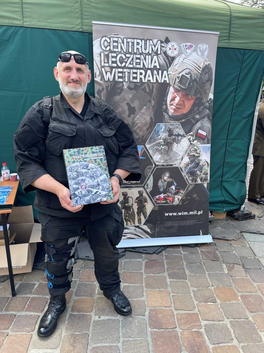 Żołnierz przy punkcie namiocie wystawienniczym Centrum Leczenia Weterana prezentuje okładkę okolicznościowej książki.