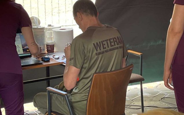 Mężczyzna podczas badań medycznych w koszulce z napisem na plecach weteran siedzi przy stoliku w namiocie wystawienniczym, obok niego pracowniczka Centrum leczenia Weterana WIM.
