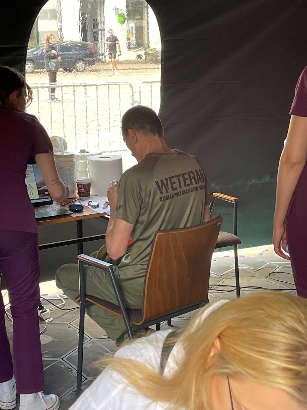 Mężczyzna podczas badań medycznych w koszulce z napisem na plecach weteran siedzi przy stoliku w namiocie wystawienniczym, obok niego pracowniczka Centrum leczenia Weterana WIM.