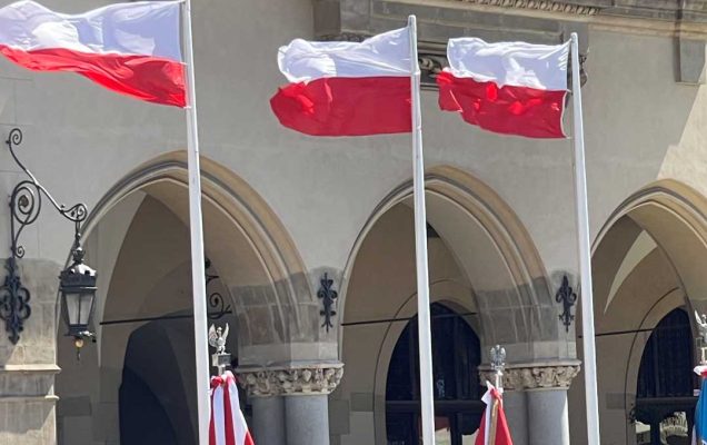 Żołnierze w strojach galowych podczas uroczystości stoją na rynku w Krakowie, z animi na masztach flagi Polski.