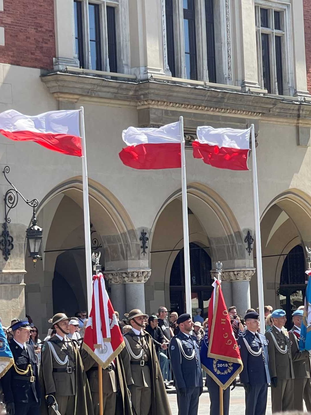 Żołnierze w strojach galowych podczas uroczystości stoją na rynku w Krakowie, z animi na masztach flagi Polski.