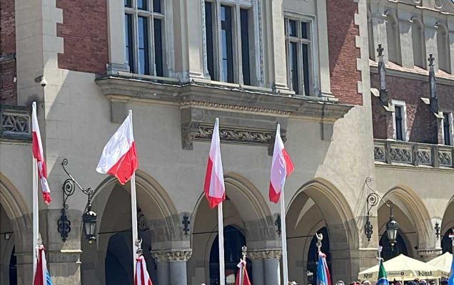 Zgromadzeni młodzi mężczyźni przyglądają się uroczystości Dnia Weterana. Przed nimi żołnierze w strojach galowych podczas uroczystości stoją na rynku w Krakowie.