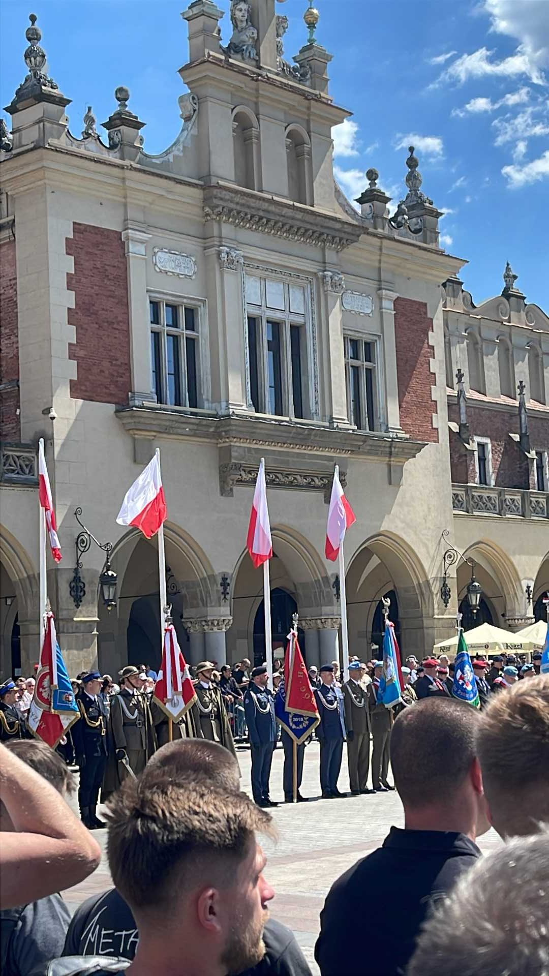 Zgromadzeni młodzi mężczyźni przyglądają się uroczystości Dnia Weterana. Przed nimi żołnierze w strojach galowych podczas uroczystości stoją na rynku w Krakowie.