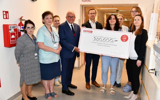 Zespół Kliniki Chorób Wewnętrznych i Hematologii WIM-PIB przedstawicie; Fundacji DKMS prezentują duży czek z wypisaną kwotą.