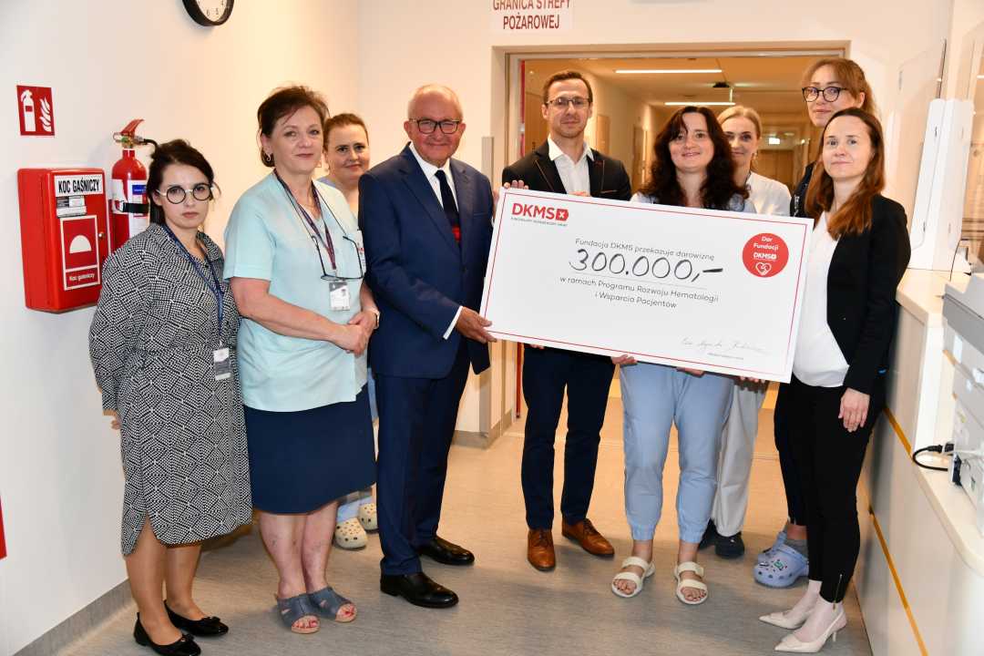 Zespół Kliniki Chorób Wewnętrznych i Hematologii WIM-PIB przedstawicie; Fundacji DKMS prezentują duży czek z wypisaną kwotą.