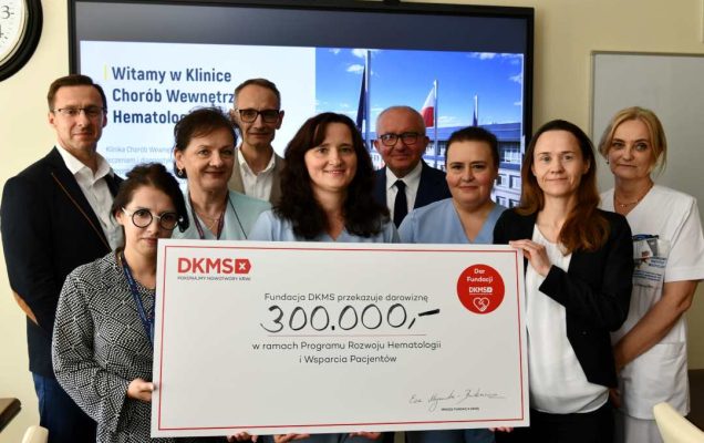 Zespół Kliniki Chorób Wewnętrznych i Hematologii WIM-PIB przedstawicie; Fundacji DKMS prezentują duży czek z wypisaną kwotą.