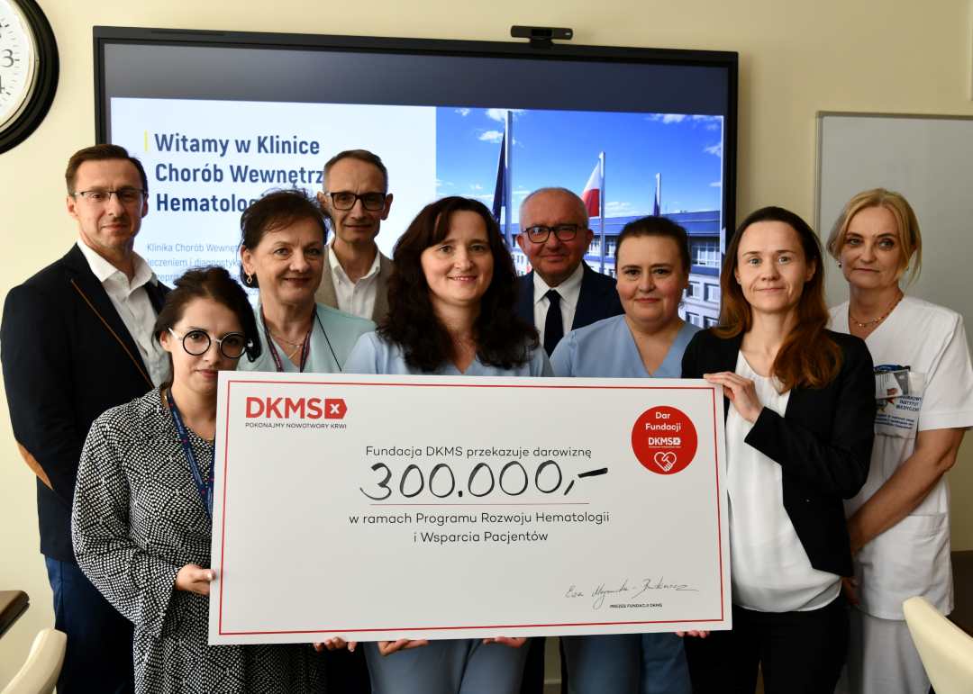 Zespół Kliniki Chorób Wewnętrznych i Hematologii WIM-PIB przedstawicie; Fundacji DKMS prezentują duży czek z wypisaną kwotą.