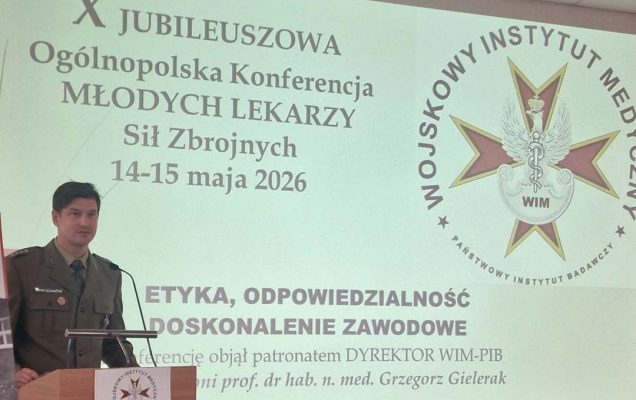 Prelegent przy mównicy za jego plecami wyświetlony slajd z konferencji.