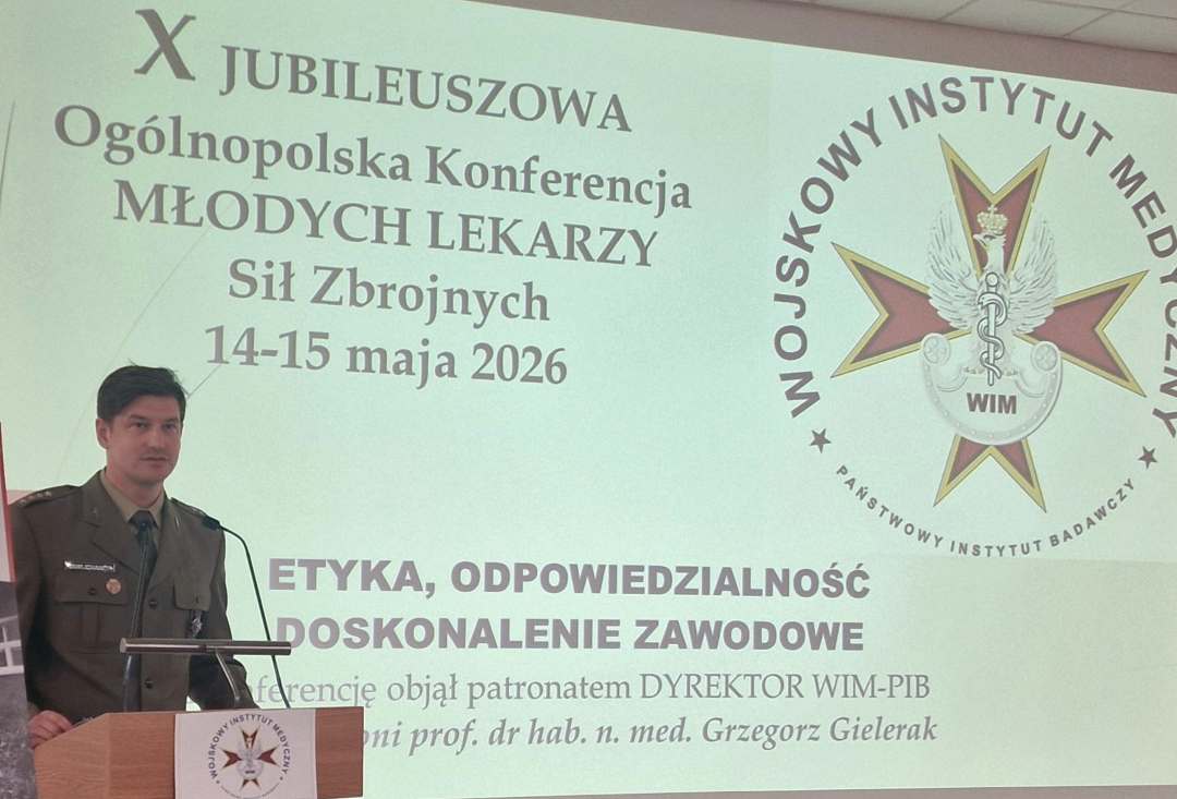 Prelegent przy mównicy za jego plecami wyświetlony slajd z konferencji.