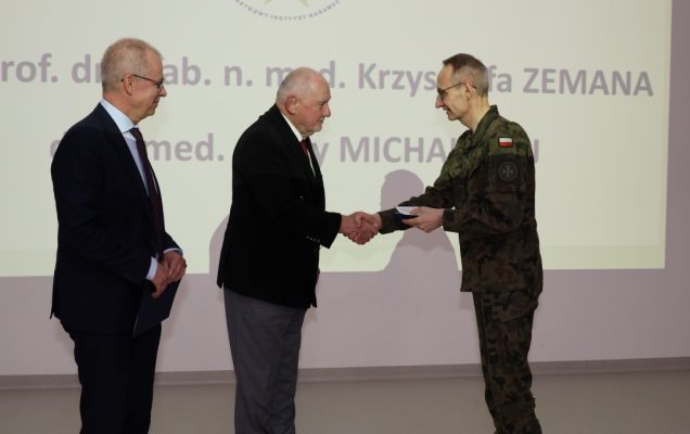Dyrektor WIM ściska dłoń prelegentowi, za nimi wyświetlony na dużym ekranie slajd z konferencji.