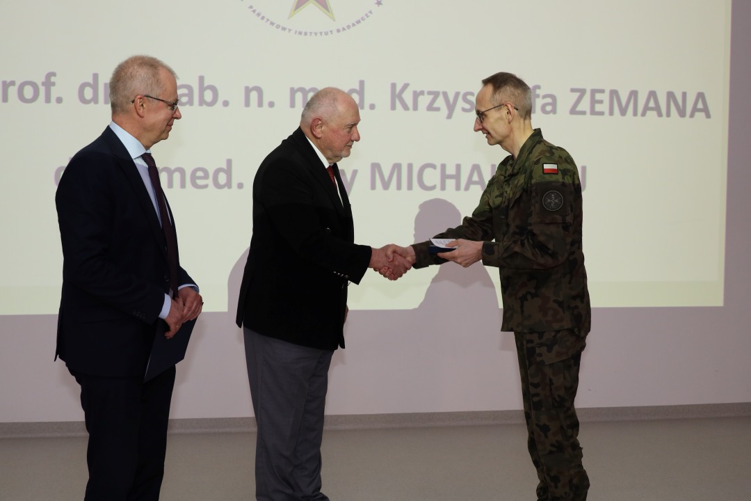 Dyrektor WIM ściska dłoń prelegentowi, za nimi wyświetlony na dużym ekranie slajd z konferencji.