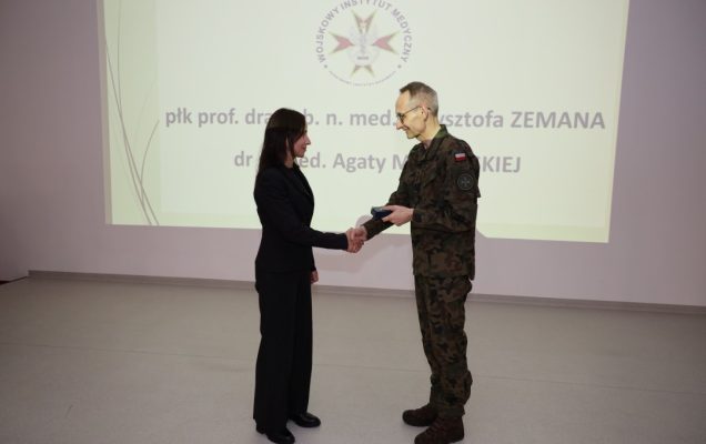 Dyrektor WIM ściska dłoń prelegentce, za nimi wyświetlony na dużym ekranie slajd z konferencji.