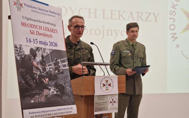 Dyrektor WIM Grzegorz Gielerak ubrany w mundur wojskowy przy mównicy otwiera konferencję. Obok niego na stojaku plakat konferencji, za jego plecami wyświetlony na ekranie slajd konferencji.