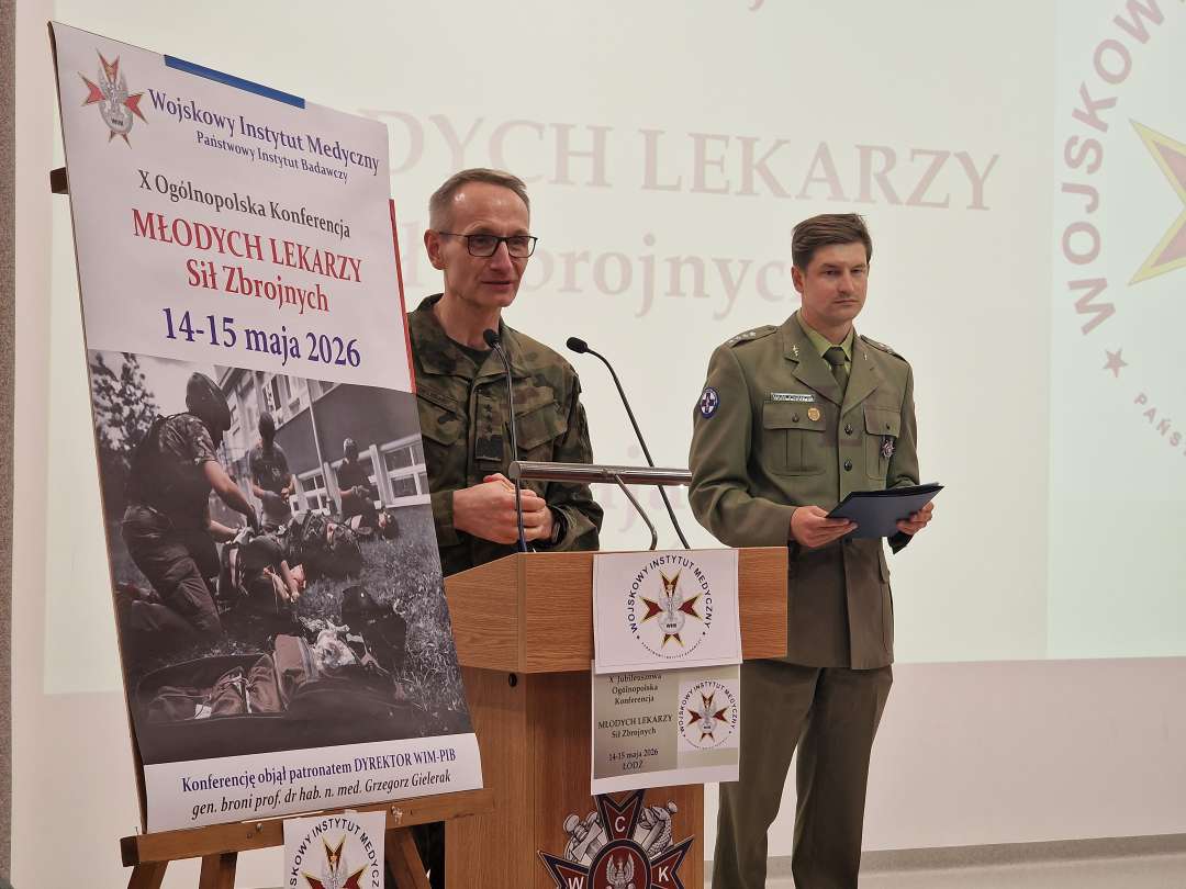 Dyrektor WIM Grzegorz Gielerak ubrany w mundur wojskowy przy mównicy otwiera konferencję. Obok niego na stojaku plakat konferencji, za jego plecami wyświetlony na ekranie slajd konferencji.