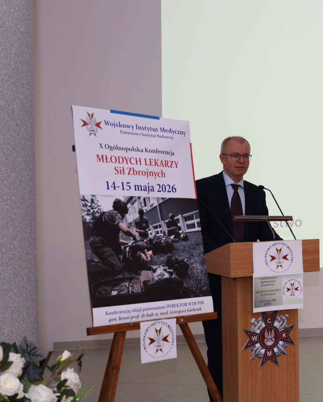 Prelegent przy mównicy, obok niego na stojaku plakat 10. konferencji Młodych Lekarzy, 14-15 maja 2026.