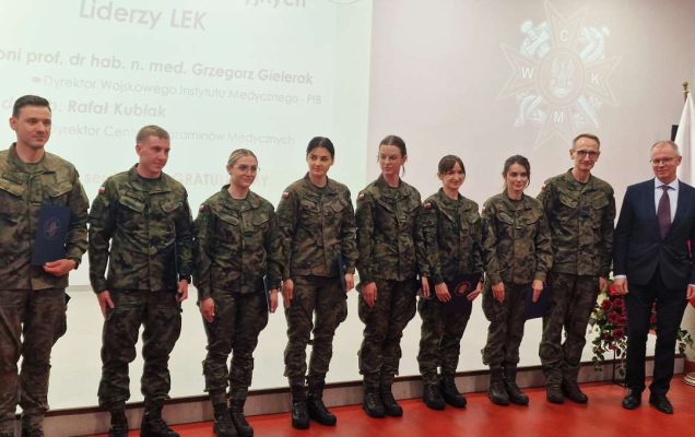 Liderzy LEK w mundurach wojskowych pozują z Dyrektorem WIM i M. Klenckim do zdjęcia na sali konferencyjnej. Za ich plecami wyświetlony slajd z konferencji.