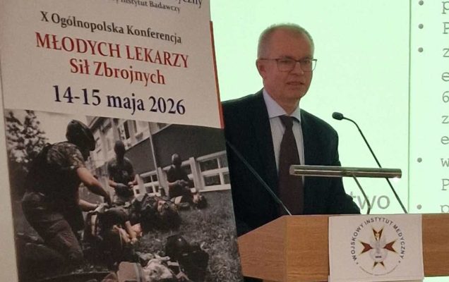Mariusz Klencki przy mównicy. Obok niego na stojaku plakat konferencyjny, za jej plecami na wielkim ekranie wyświetlony slajd z prezentacji.