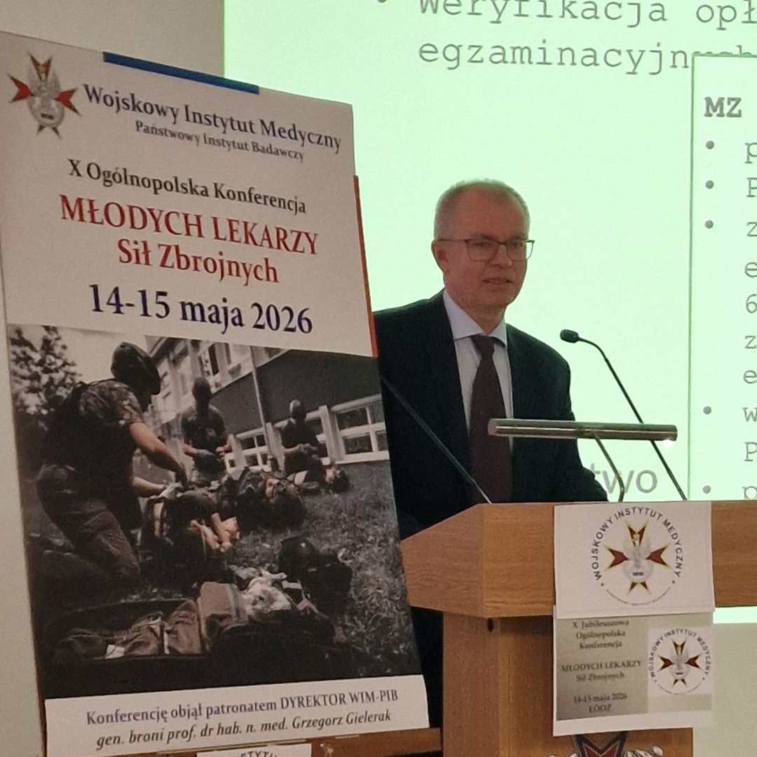Mariusz Klencki przy mównicy. Obok niego na stojaku plakat konferencyjny, za jej plecami na wielkim ekranie wyświetlony slajd z prezentacji.