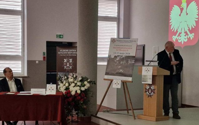 Profesor Zeman na sali podczas wykładu. Obok niego na stojaku plakat konferencyjny, za jego plecami godło RP.