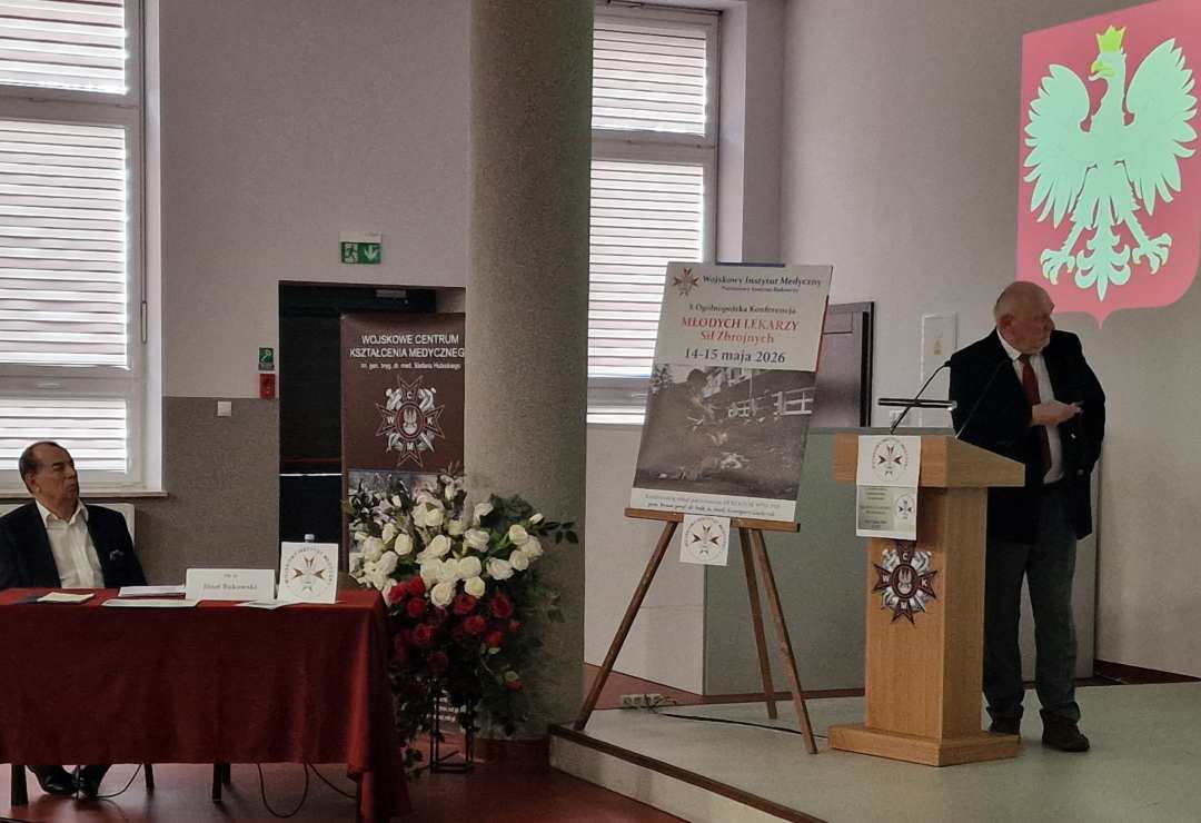 Profesor Zeman na sali podczas wykładu. Obok niego na stojaku plakat konferencyjny, za jego plecami godło RP.