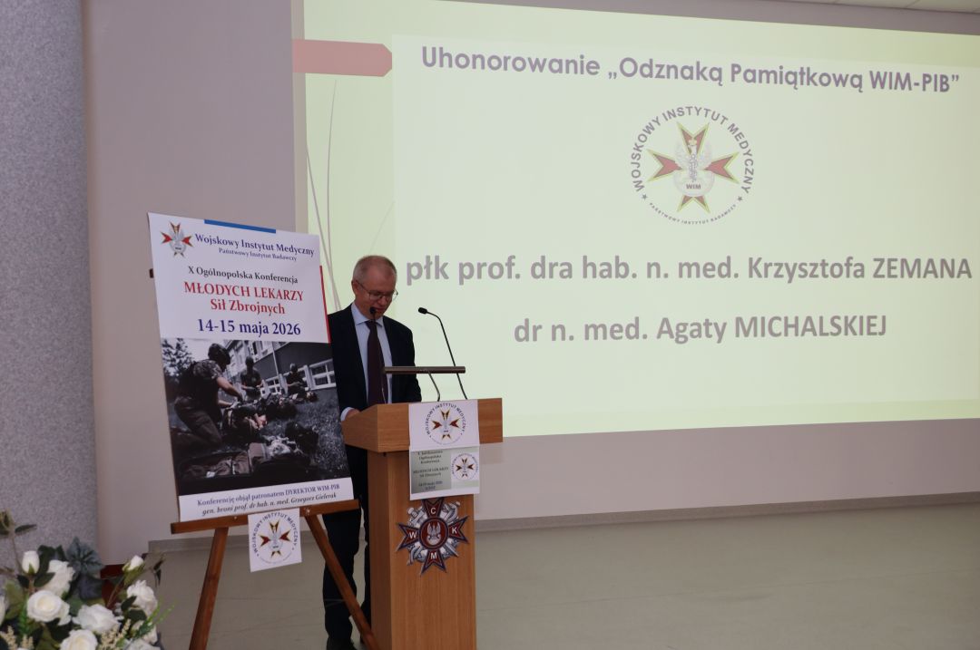 Prelegent przy mównicy, za jego plecami na ekranie slajd z konferencji.
