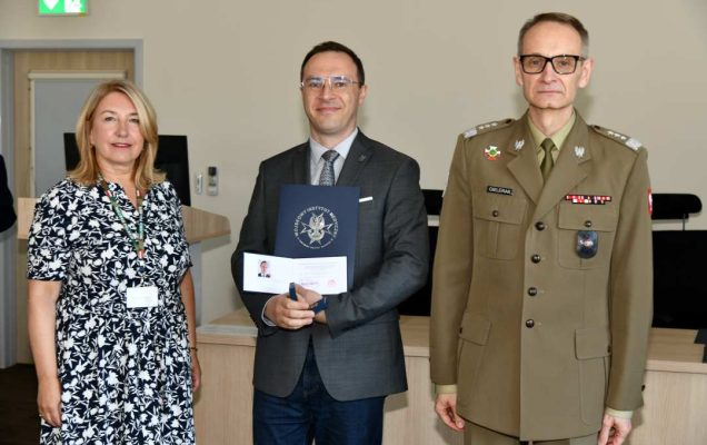 Grzegorz Gielerak i Marta Maciążek pozują z absolwentem stojącym pomiędzy nimi. Mężczyzna trzyma w rękach dyplom ukończenia studiów oraz otwarty drugi duplom z wyróżnieniem.