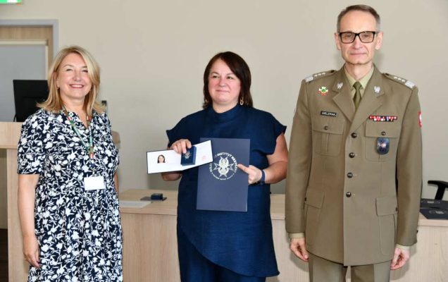 Grzegorz Gielerak i Marta Maciążek pozują z absolwentką stojącą pomiędzy nimi. Kobieta trzyma w jedej ręce dyplom w drugiej ręce prezentuje dyplom z wyróżnieniem i granatowe pudełko ze schowaną odznaką.
