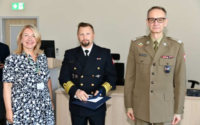 Grzegorz Gielerak i Marta Maciążek pozują z absolwentem stojącym pomiędzy nimi. Mężczyzna jest ubrany w granatowy mundur, trzyma w rękach dyplom ukończenia studiów.