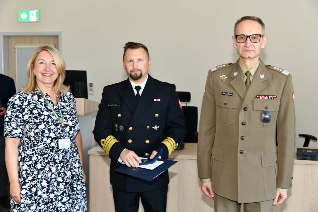 Grzegorz Gielerak i Marta Maciążek pozują z absolwentem stojącym pomiędzy nimi. Mężczyzna jest ubrany w granatowy mundur, trzyma w rękach dyplom ukończenia studiów.
