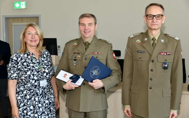 Grzegorz Gielerak i Marta Maciążek pozują z absolwentem stojącym pomiędzy nimi. Mężczyzna jest ubrany w mundur wojskowy, trzyma w rękach dyplom ukończenia studiów oraz otwarty drugi duplom z wyróżnieniem.