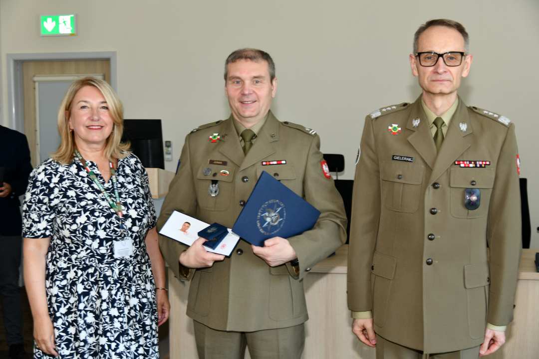 Grzegorz Gielerak i Marta Maciążek pozują z absolwentem stojącym pomiędzy nimi. Mężczyzna jest ubrany w mundur wojskowy, trzyma w rękach dyplom ukończenia studiów oraz otwarty drugi duplom z wyróżnieniem.