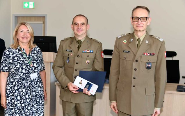 Grzegorz Gielerak i Marta Maciążek pozują z absolwentem stojącym pomiędzy nimi. Mężczyzna ubrany jest w mundur wojskowy, w rękach dyplom ukończenia studiów oraz otwarty drugi duplom z wyróżnieniem.