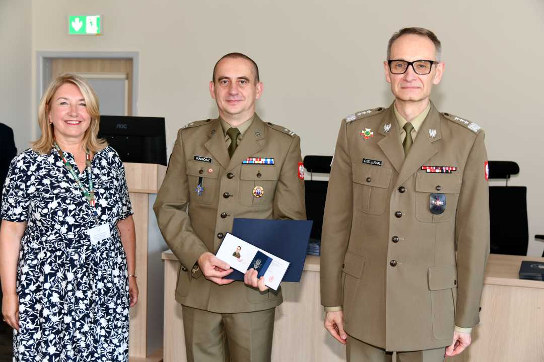 Grzegorz Gielerak i Marta Maciążek pozują z absolwentem stojącym pomiędzy nimi. Mężczyzna ubrany jest w mundur wojskowy, w rękach dyplom ukończenia studiów oraz otwarty drugi duplom z wyróżnieniem.