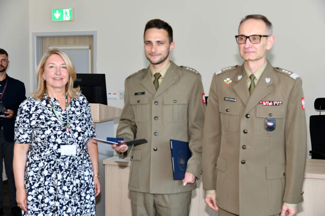 Grzegorz Gielerak i Marta Maciążek pozują z absolwentem stojącym pomiędzy nimi. Mężczyzna ubrany jest w mundur wojskowy, w rękach dyplom ukończenia studiów.
